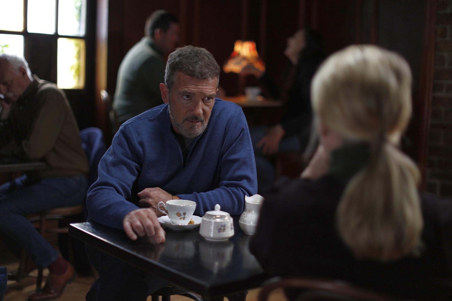 Ein Mann in blauem Pullover sitzt an einem Tisch mit einer Teetasse, einer Frau mit blondem Pferdeschwanz gegenüber. Andere Personen im Hintergrund.