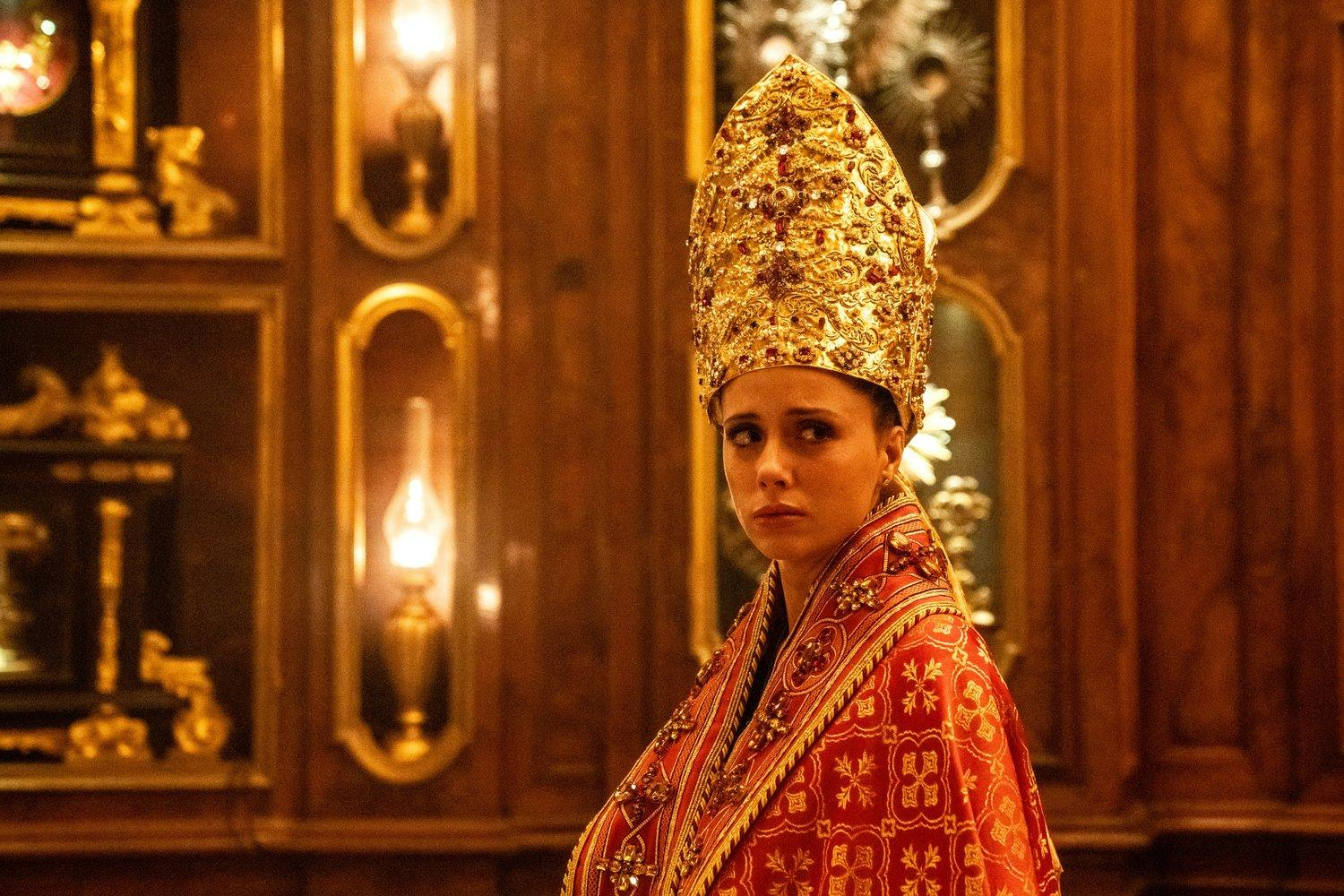 Eine Person mit goldener Krone und rotem Gewand steht in einer Kirche. Der Hintergrund hat Holzpaneele und Lichter. Die Person wirkt besorgt.
