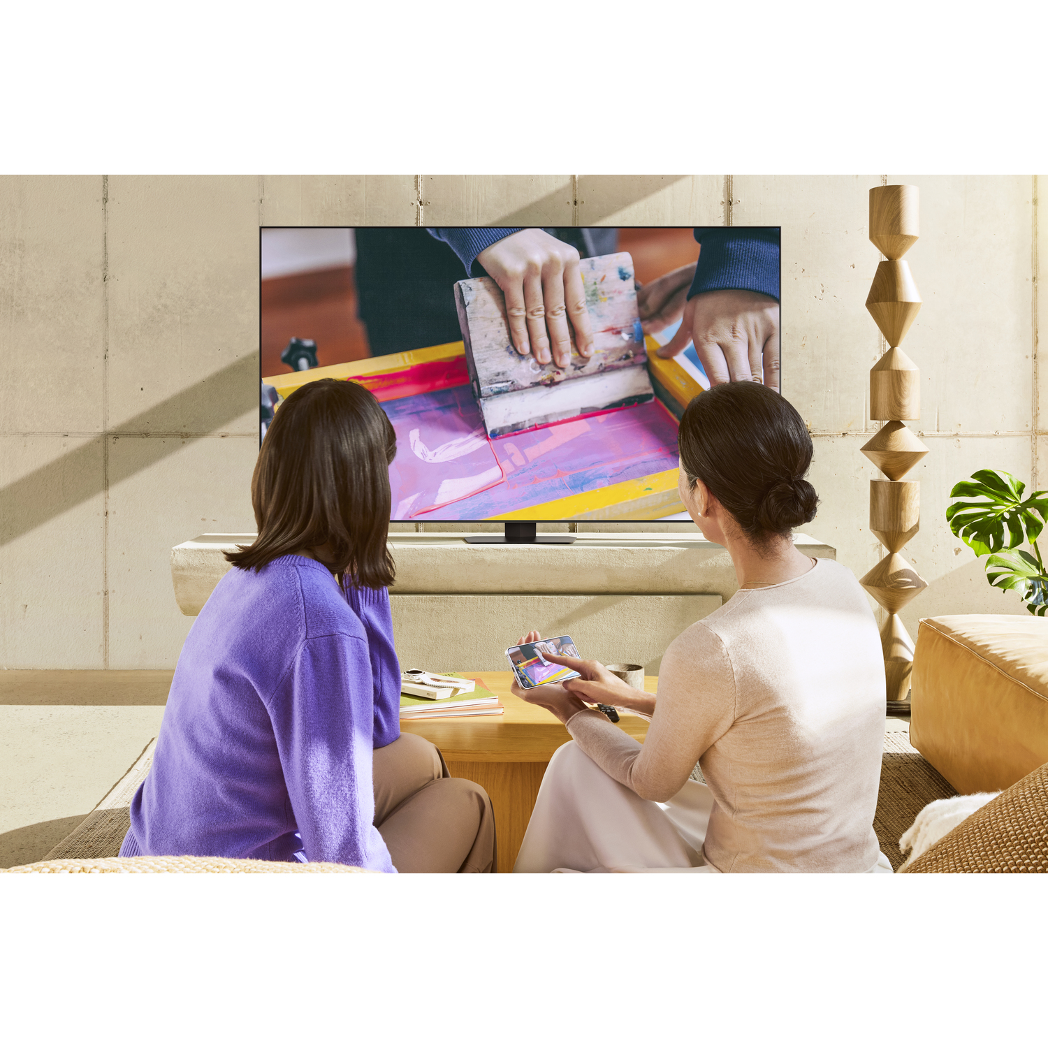 Twee vrouwen zitten voor een tv die een persoon toont die met een gereedschap werkt.