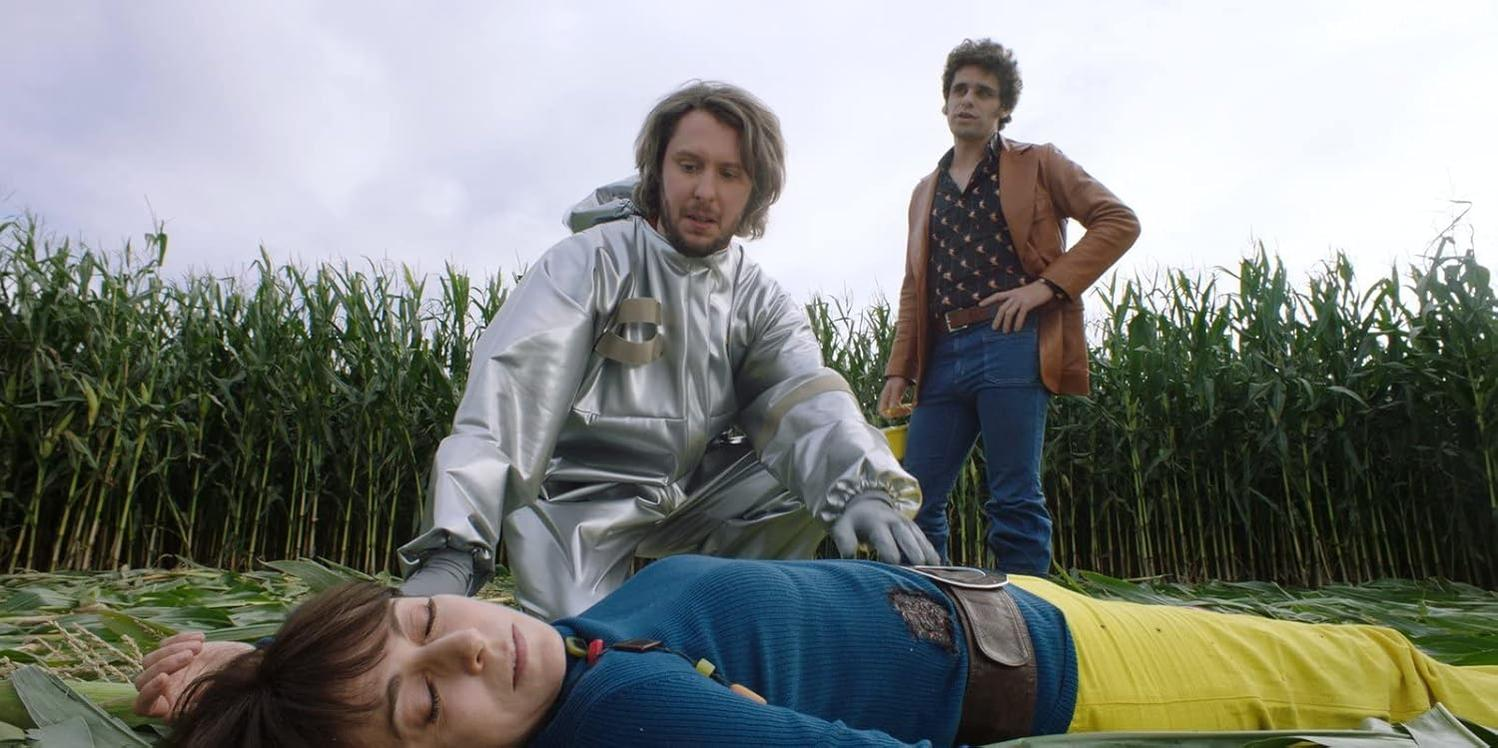 Ein Mann in Silber neben einem Mann, der auf einem Feld liegt. Ein weiterer Mann steht daneben.