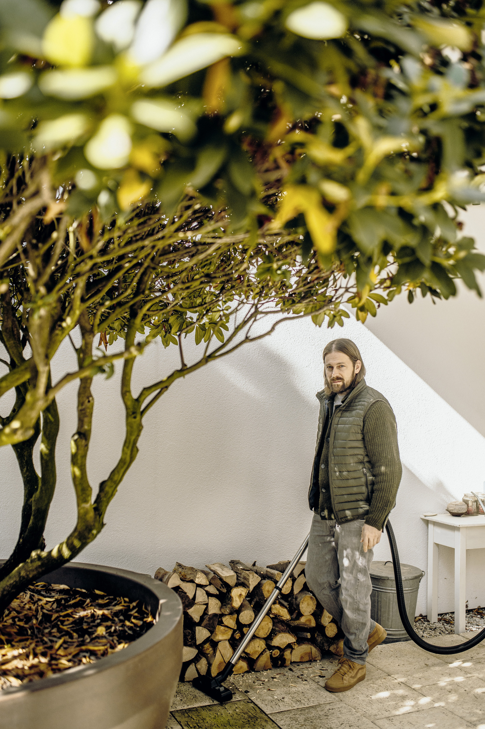 Mężczyzna odkurza na zewnątrz pod drzewem. Ma na sobie kamizelkę i dżinsy. Jest stos drewna.
