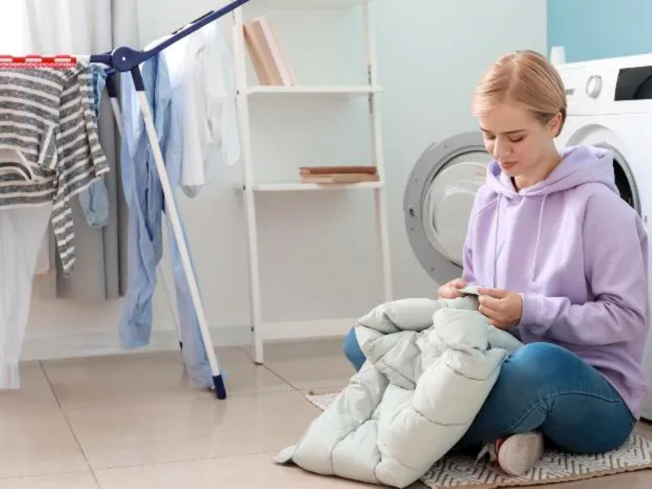 Kobieta siedzi na podłodze w pralni, szyjąc kurtkę. Ubrania wiszą w tle.