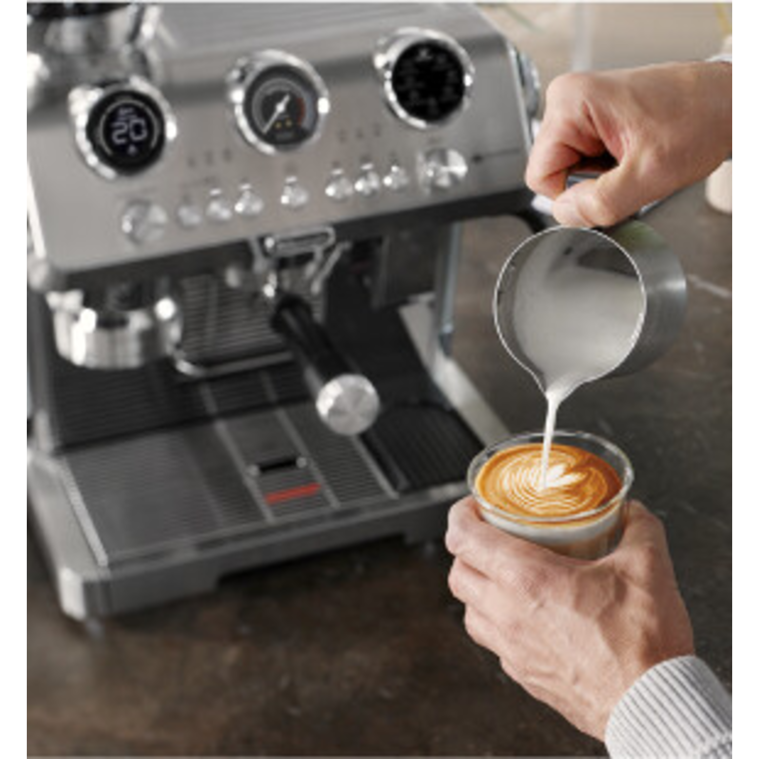 Hands pouring milk into a glass of coffee. A coffee machine is visible in the background.