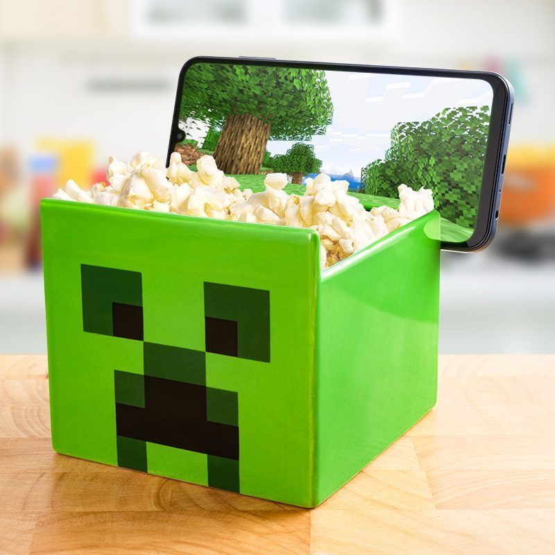 Zöld Creeper pattogatott kukorica tartály pattogatott kukoricával. Egy telefon Minecraft jelenetet mutat. Fa asztalon.