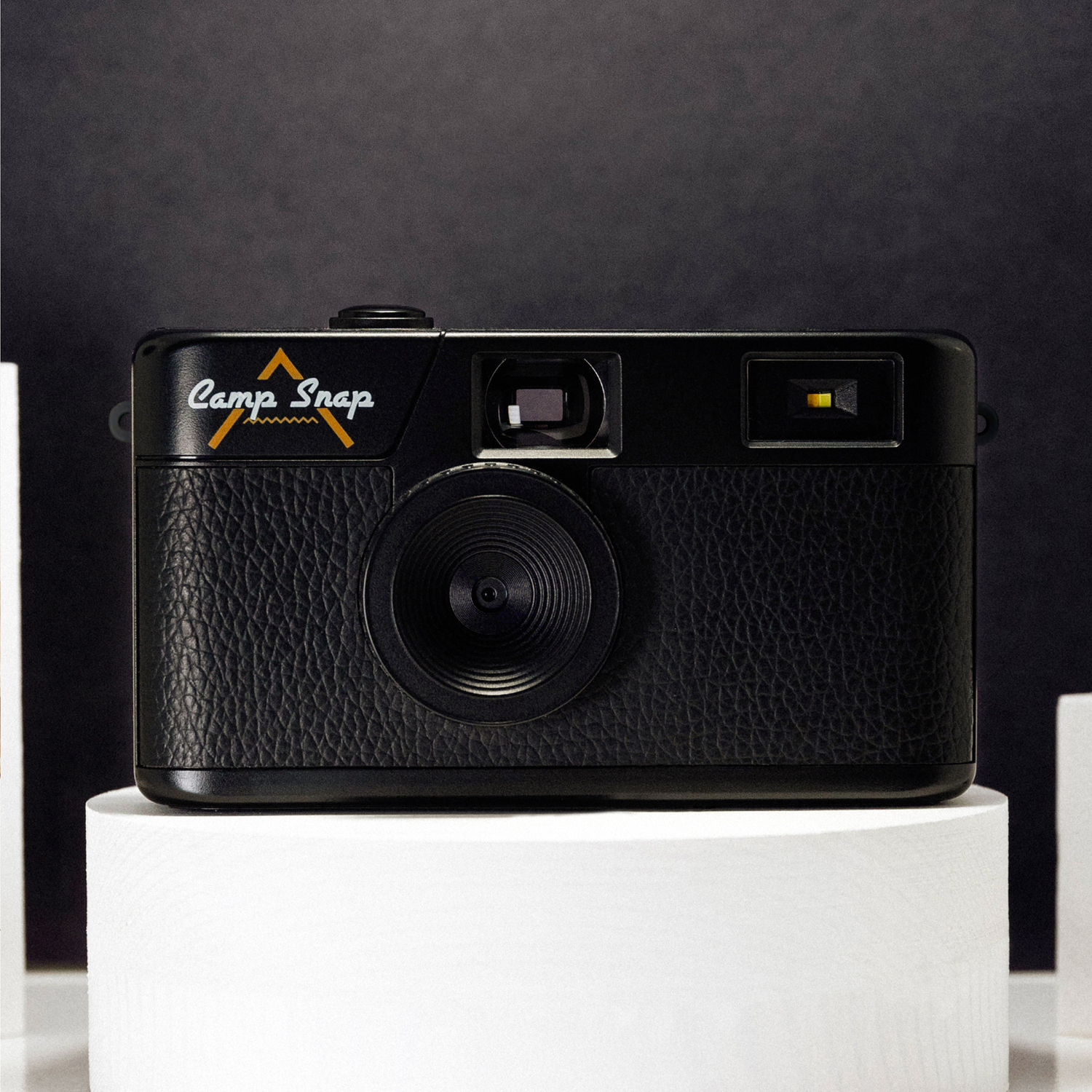 Zwarte Camp Snap camera op een witte standaard tegen een donkere achtergrond. De camera heeft een getextureerde grip en een prominente lens.