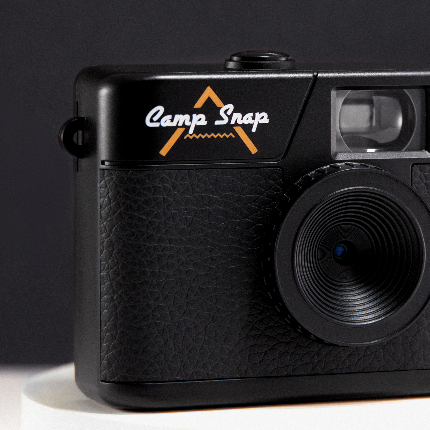 Close-up van een zwarte camera met de tekst 'Camp Snap'. De camera heeft een getextureerde grip en een lens. De achtergrond is donker.
