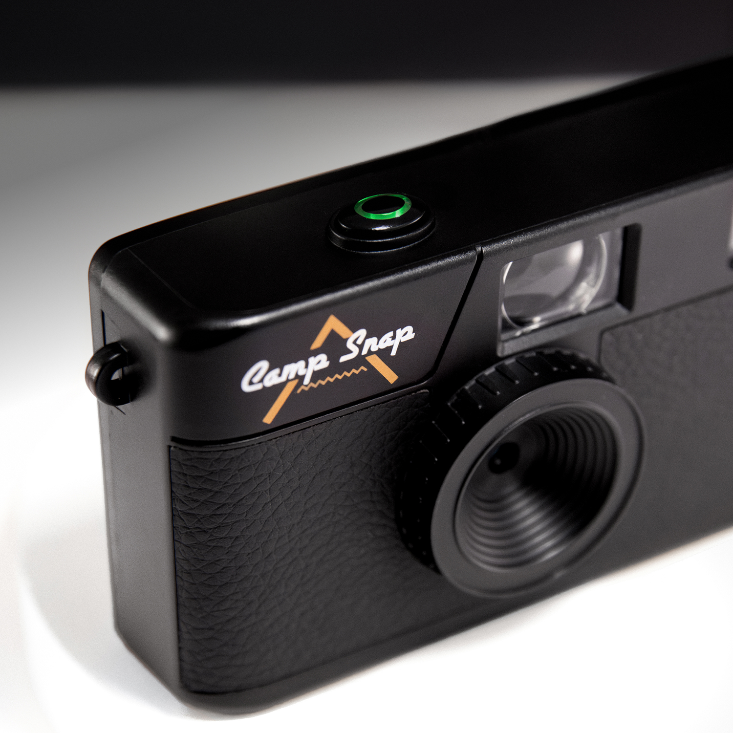 Zwarte Camp Snap camera. Groene knop, gouden logo en lens detail. Achtergrond is wit en zwart.