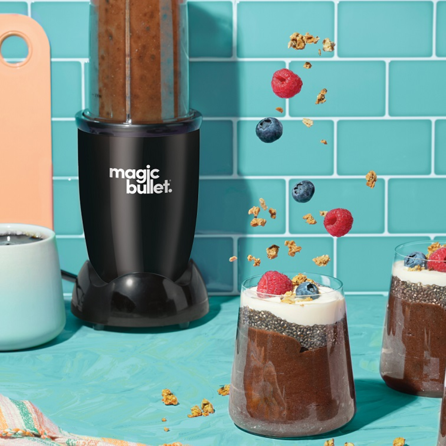 Black Magic Bullet blender with chocolate smoothie, berries, and granola. Glasses with smoothie, berries, and granola. Turquoise background.