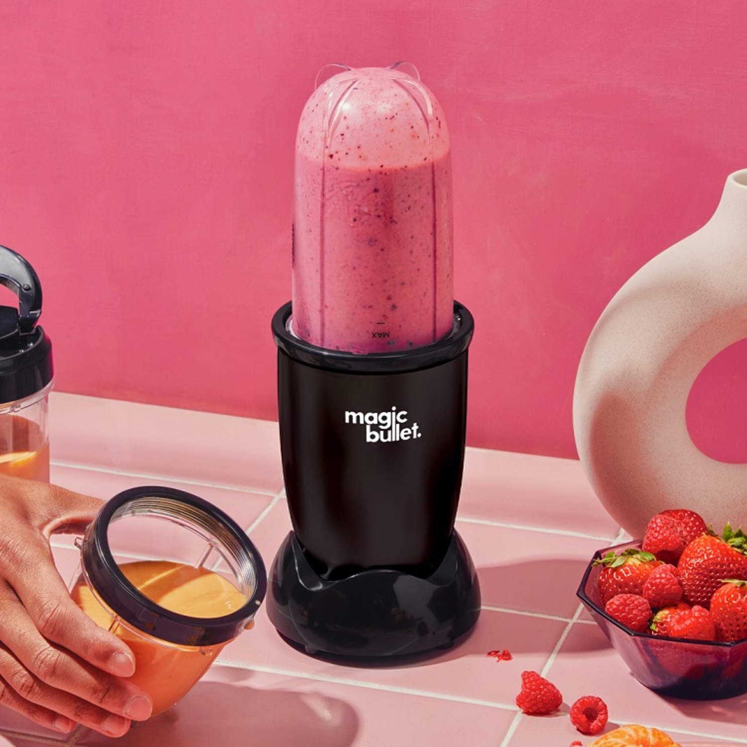 Black Magic Bullet blender with pink smoothie, a glass with smoothie, and a bowl of strawberries on pink tiles.