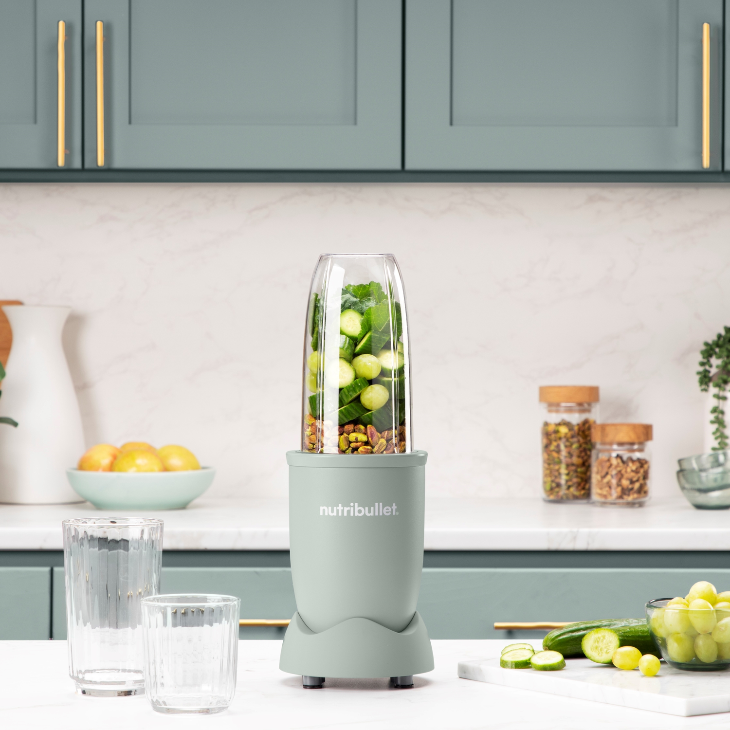 A green Nutribullet blender with cucumbers and pistachios. On a kitchen countertop with glasses and vegetables.