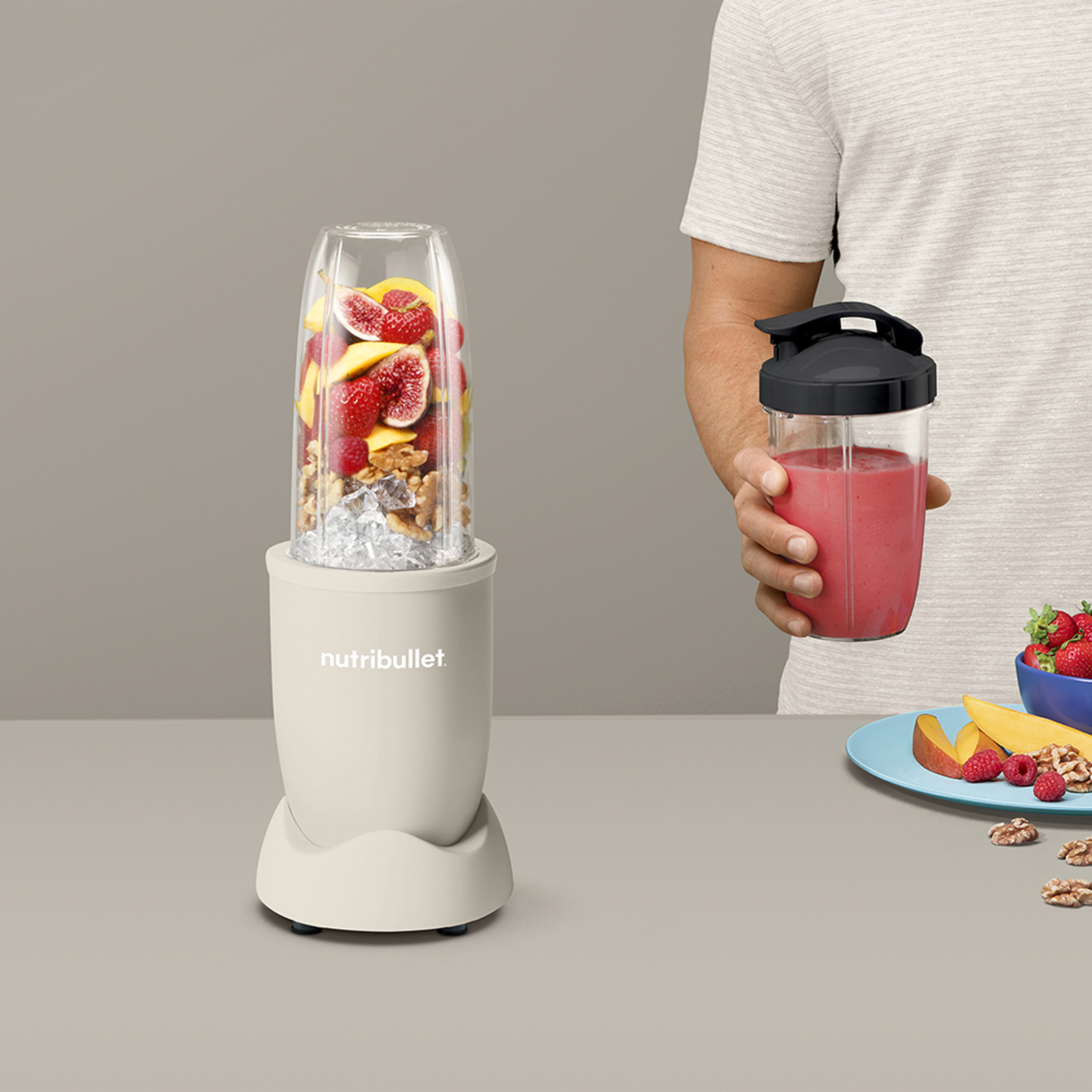 Blender filled with fruits and nuts. A person holds a smoothie cup, fruits beside it.