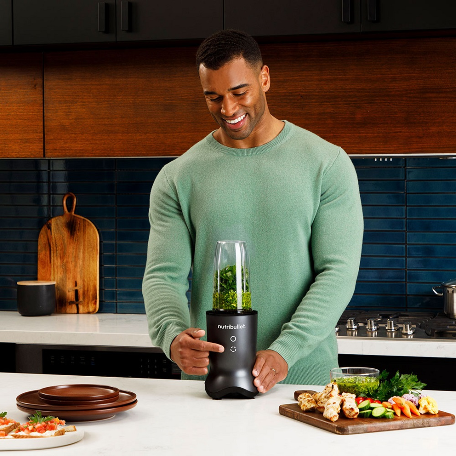 A man holding a black blender and pressing a button. Ingredients for a dish on the table.