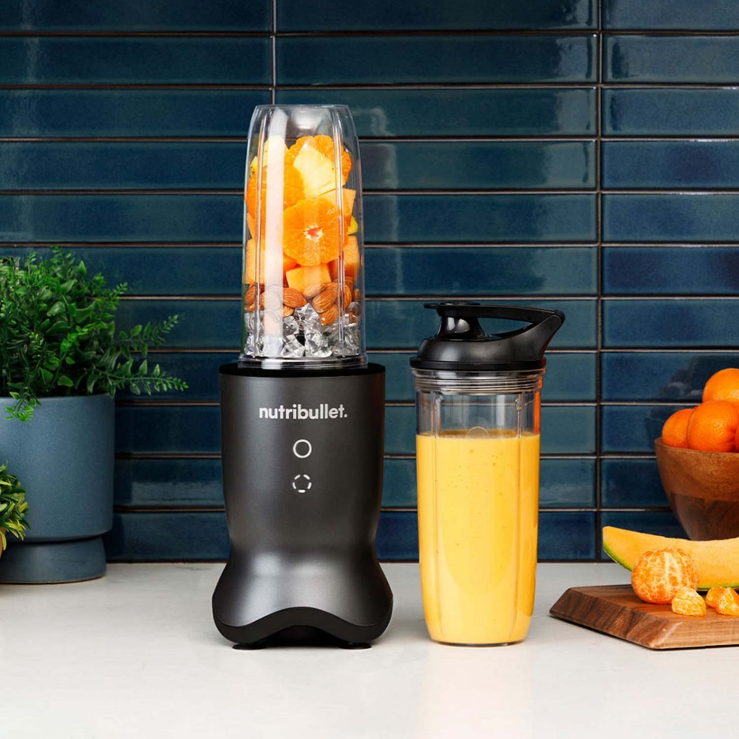 Black blender with tangerines and almonds. A smoothie cup with yellow content next to it.