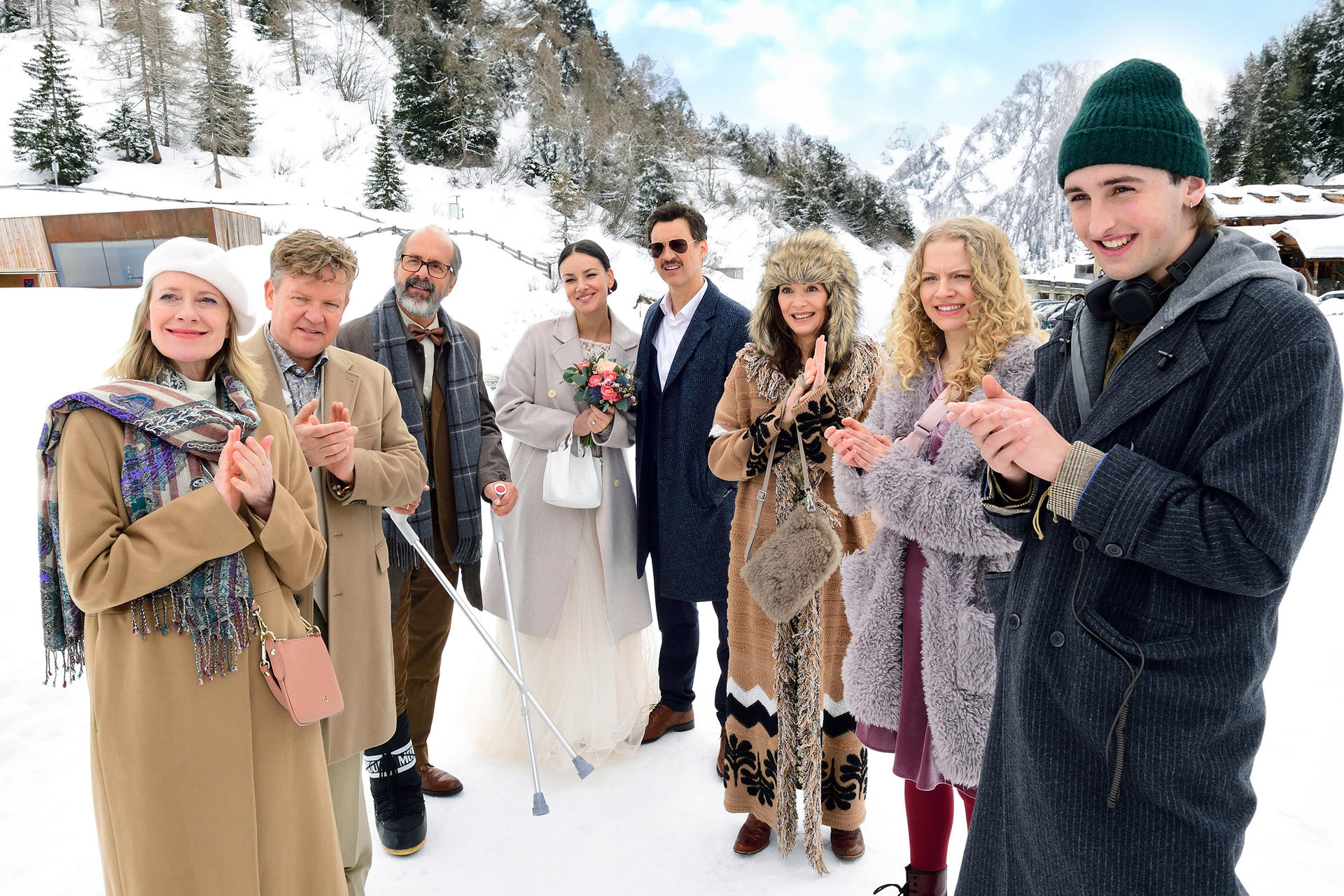Gruppe von Menschen im Schnee, einige klatschen, im Hintergrund Berge und Gebäude.