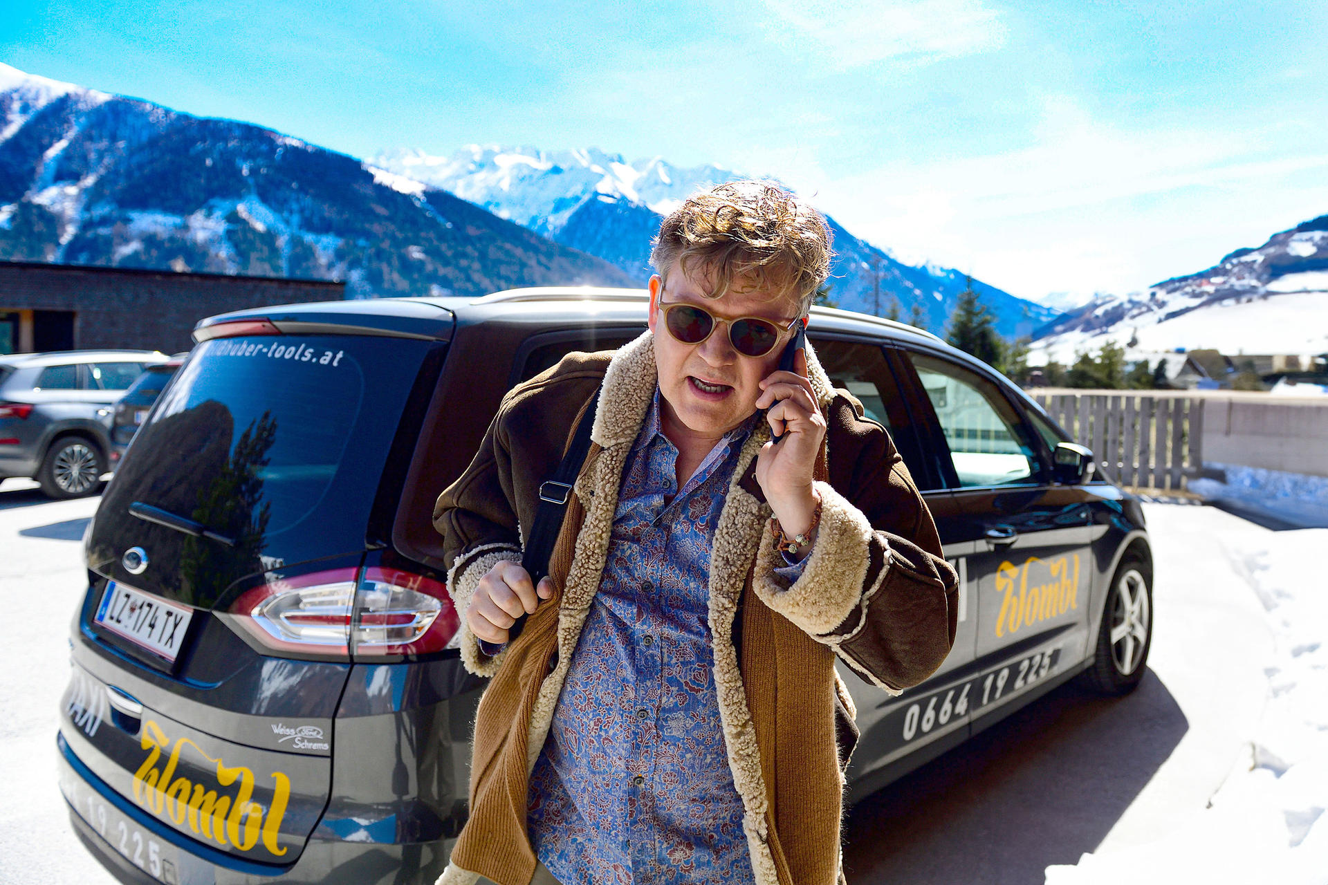 Mann mit Handy vor einem Auto mit Schneebergen im Hintergrund.