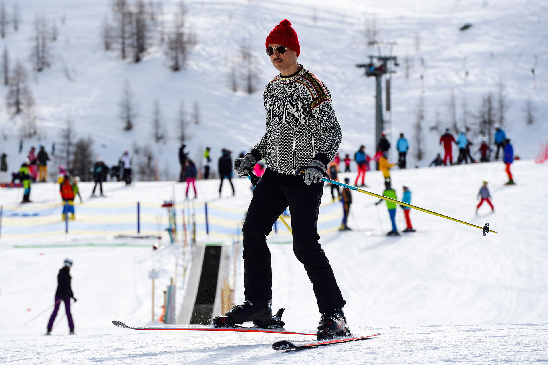 Ein Mann in einem roten Hut und Pullover fährt Ski mit anderen Skifahrern im Hintergrund.
