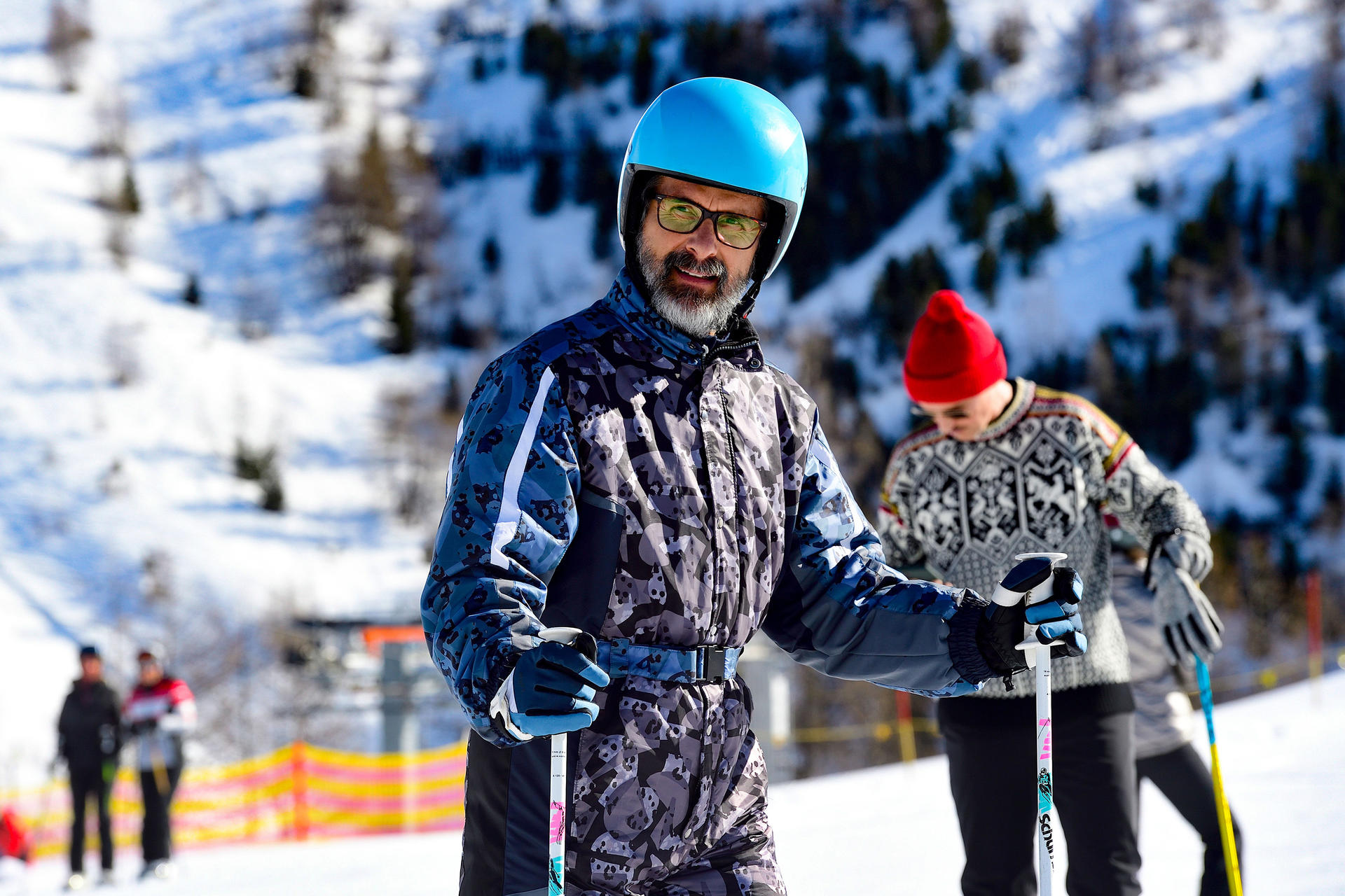 Ein Mann mit blauem Helm und Skianzug steht im Schnee, hält Skistöcke.