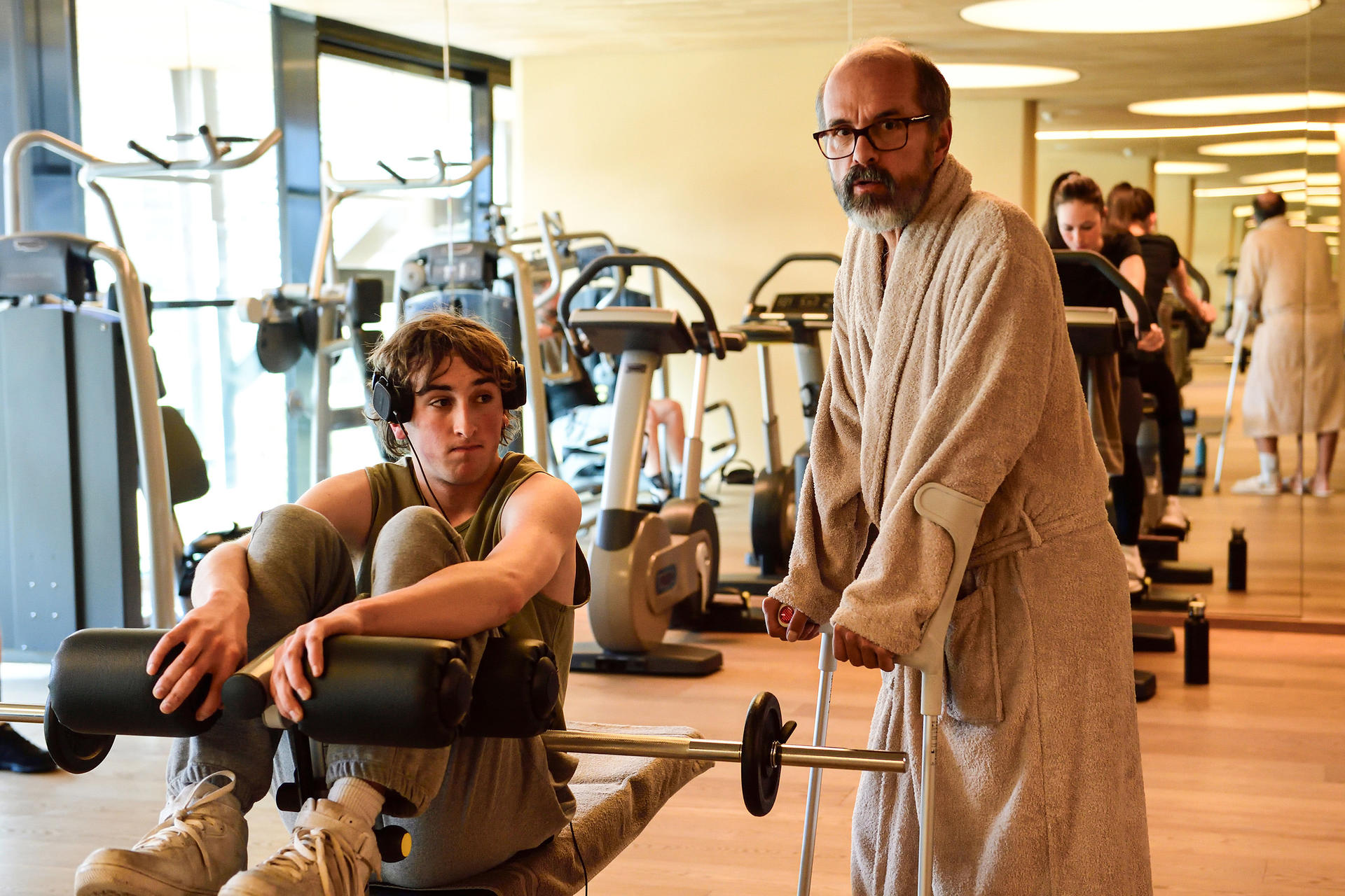 Ein Mann mit Krücken und Bademantel steht neben einem Jungen mit Kopfhörern im Fitnessstudio.