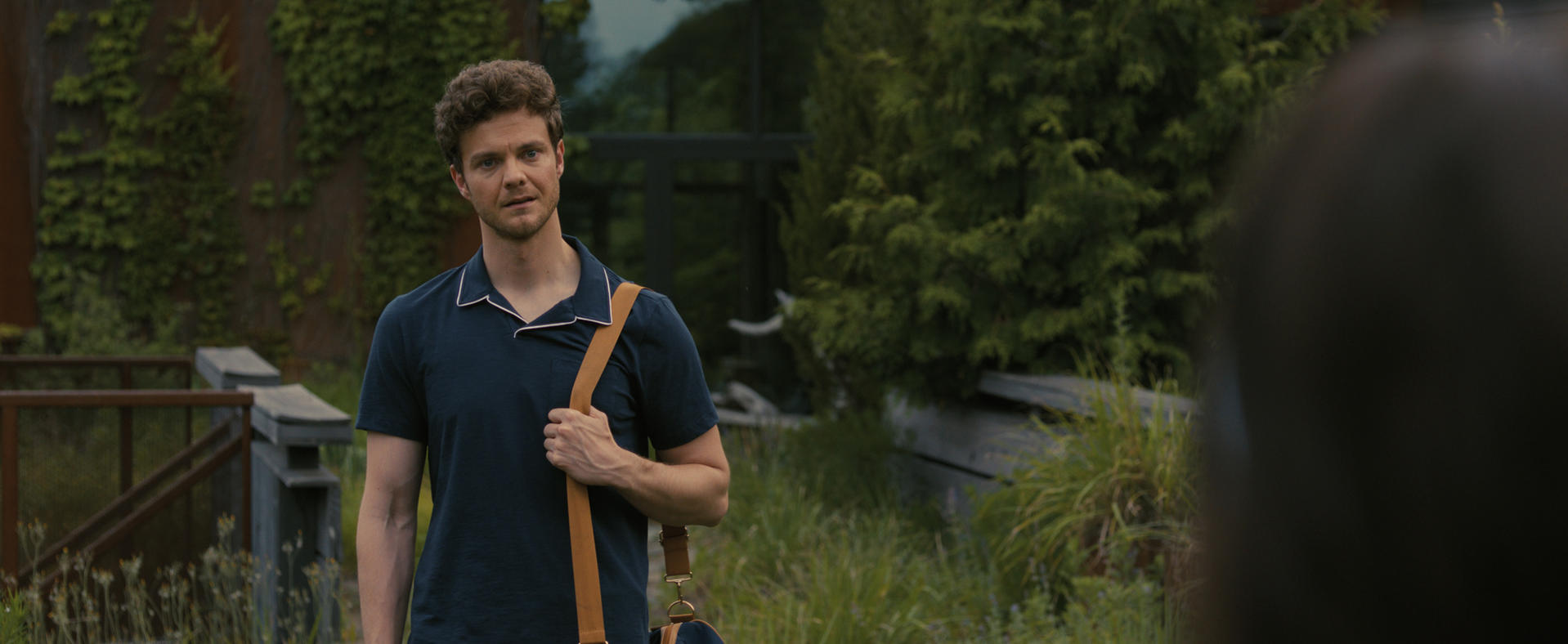 Ein Mann mit lockigem Haar in einem blauen Poloshirt mit braunem Gurt steht draußen.