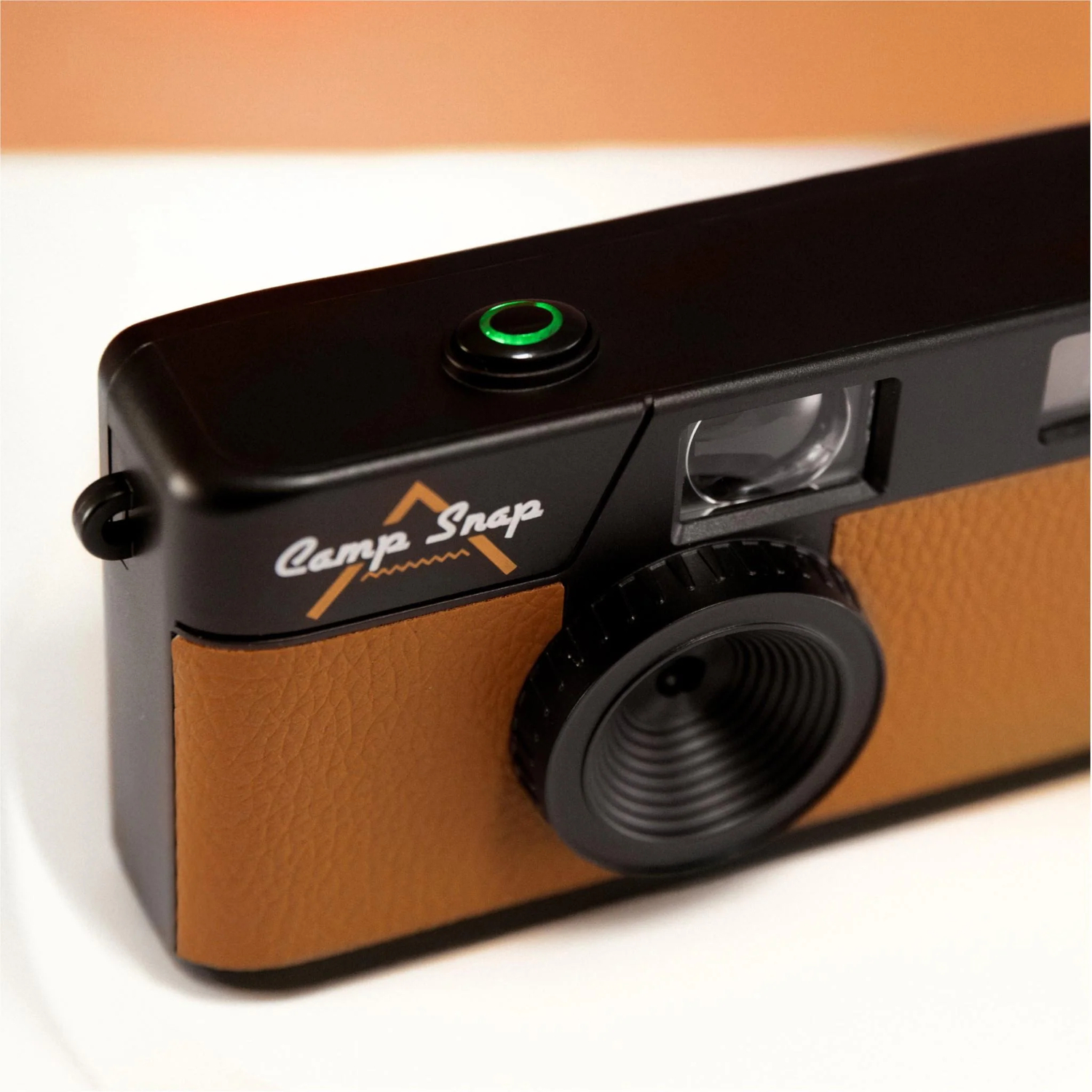 A close-up of a black and tan camera with the words 'Camp Snap' on it. The button is green.