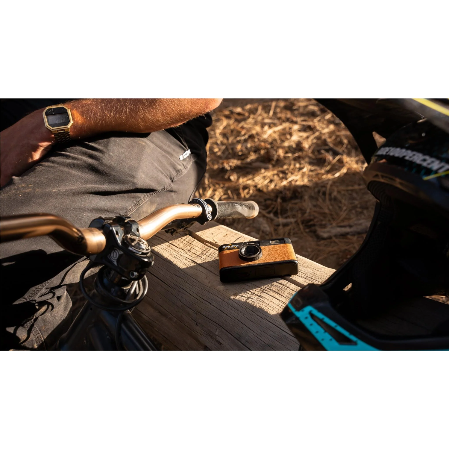A person with a gold watch sits next to a bike. A camera sits on a wooden surface next to a helmet.