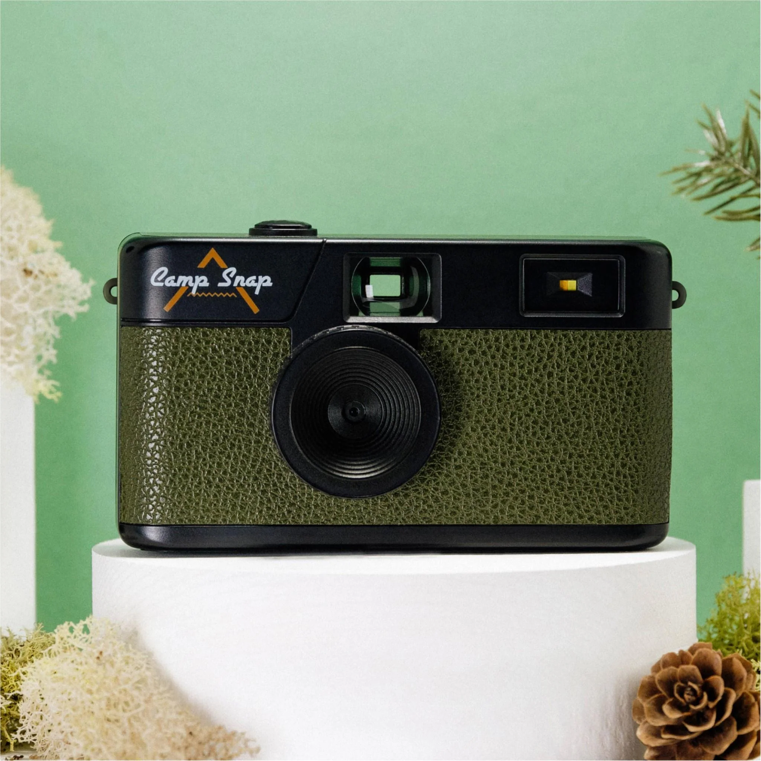 A green and black Camp Snap camera sits on a white pedestal. The background is green with pine and moss accents.