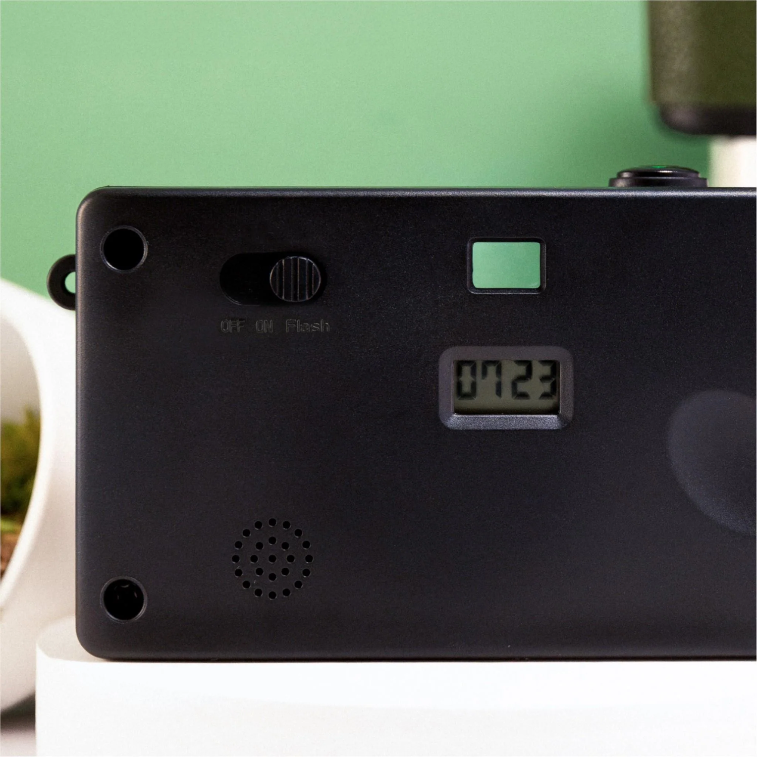 Black camera back with a display showing '0723'. It has a flash switch, green elements, and a speaker. Background is green.