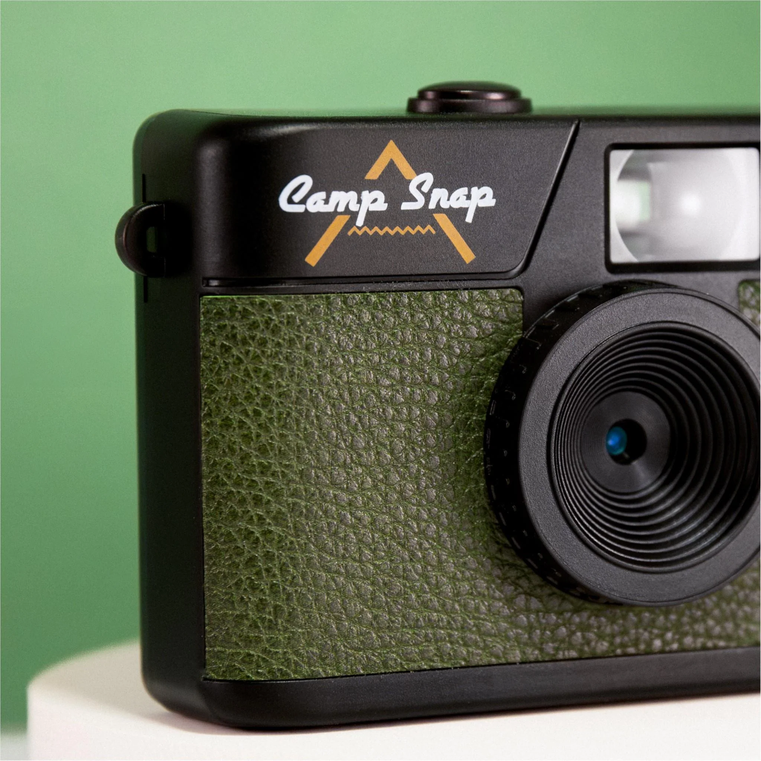Black camera with green leather. 'Camp Snap' logo. Lens, flash, and details visible. Background is green.