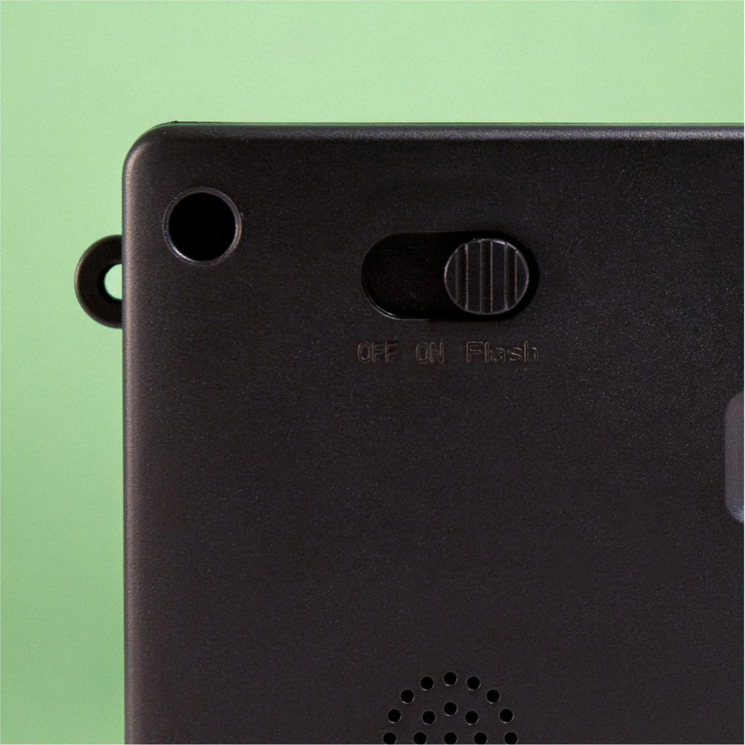 Close-up of a black device. Features an on/off/flash switch, a small hole, and a loop. Green background.