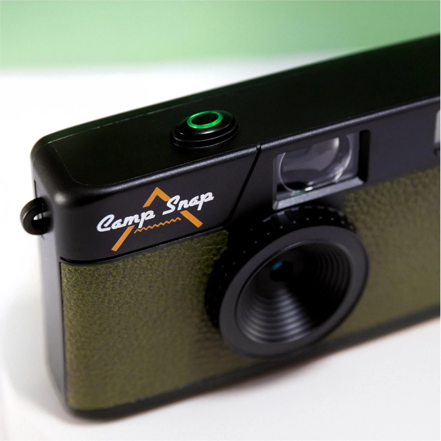 A close-up of a black and olive green camera with the words 'Camp Snap' on it. The camera has a lens and a green button.