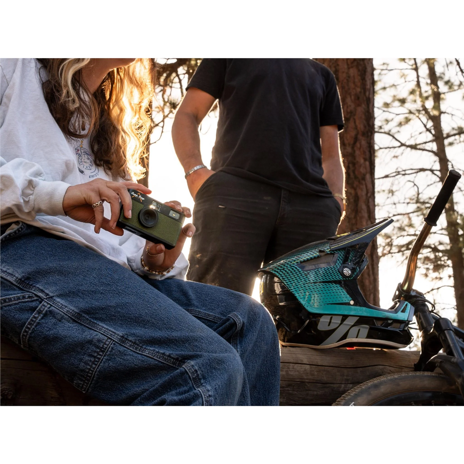 A person holds a green camera, and another stands nearby. A helmet rests on a log, next to a bicycle and trees in the background.