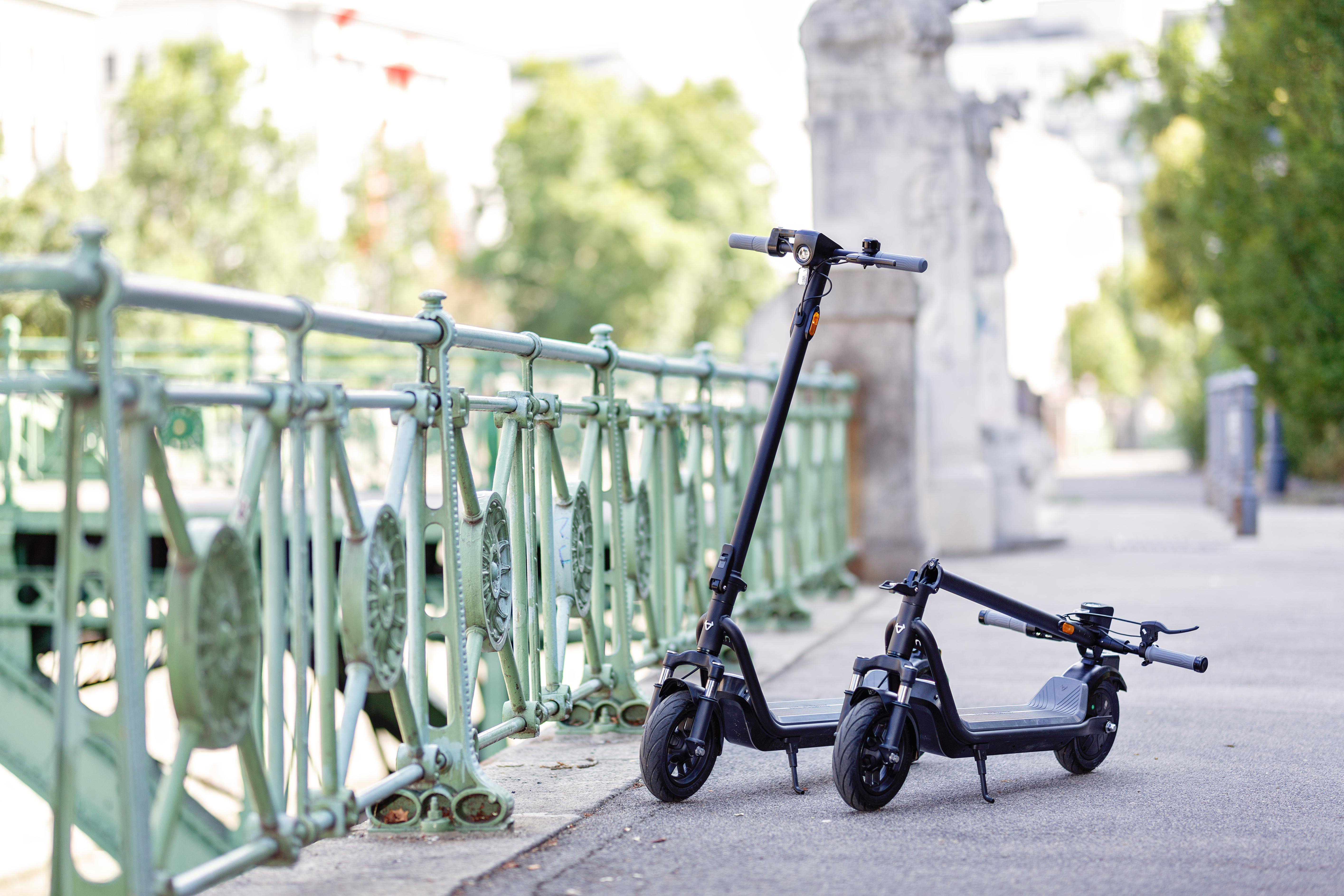 Zwei schwarze Elektroroller stehen auf einem Gehweg neben einer Brücke. Der Hintergrund ist verschwommen.