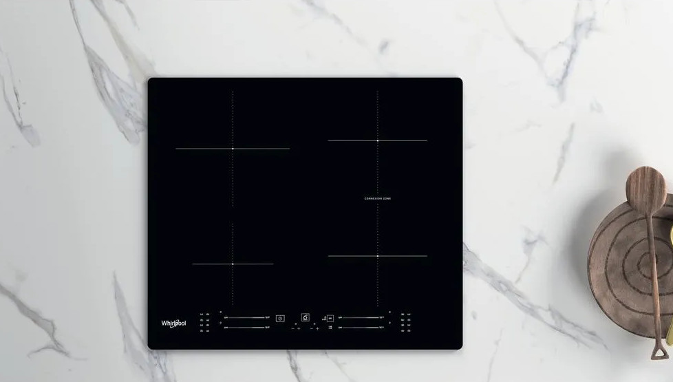 Czarna płyta indukcyjna z czterema palnikami na marmurowej powierzchni. Drewniana łyżka i talerz obok.