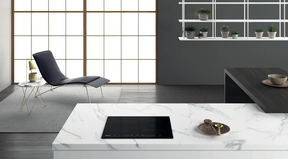 Nowoczesna scena kuchenna. Czarna płyta grzewcza na marmurowym blacie. Krzesło i stół przy oknie. Rośliny na półkach. Szara ściana.