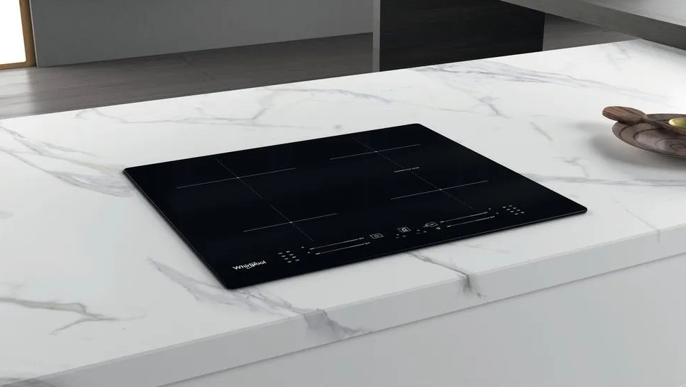 Czarna płyta indukcyjna Whirlpool na białym marmurowym blacie. Drewniana miska w tle.