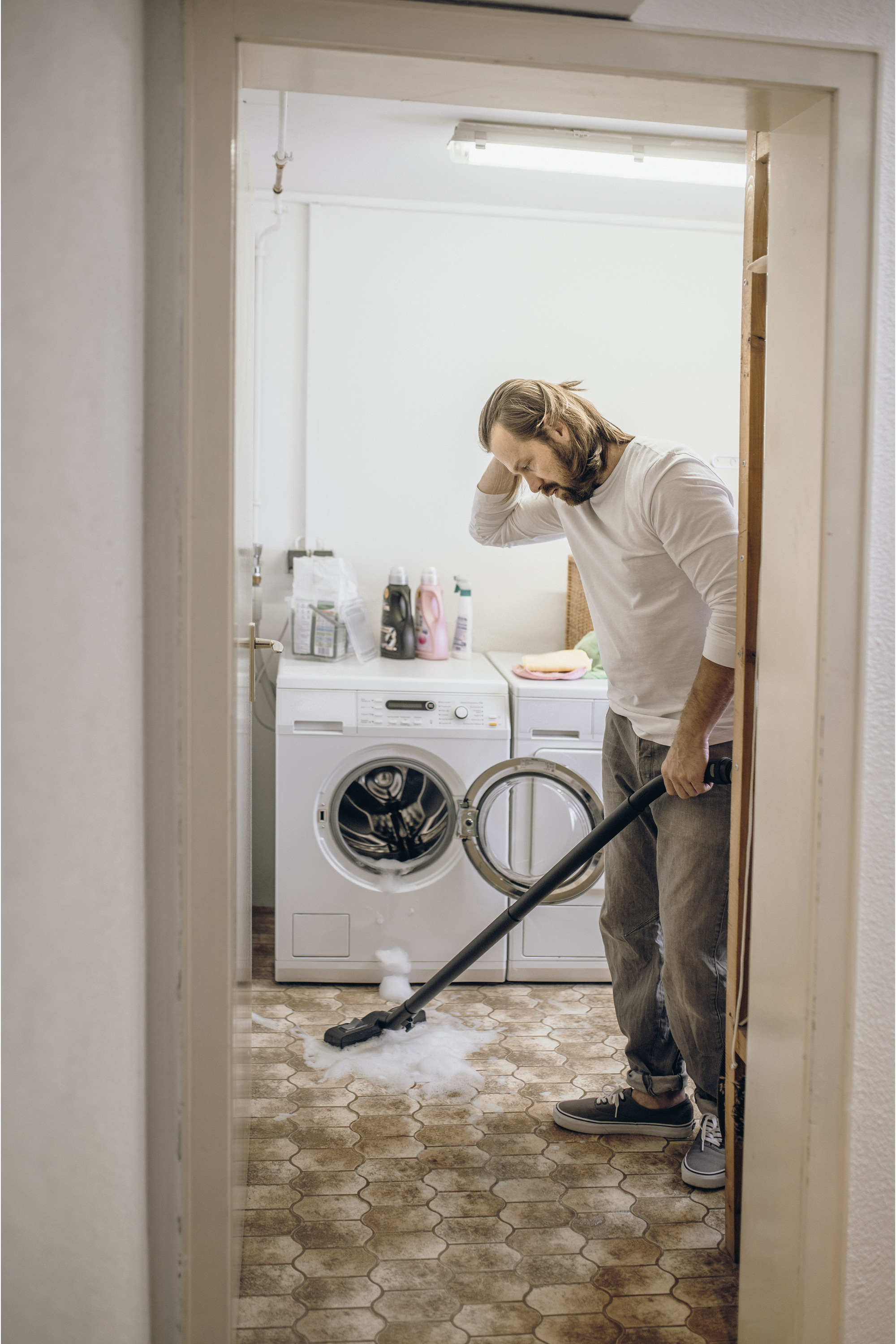 Mężczyzna odkurza w pralni. Ma na sobie dżinsy i białą koszulę.