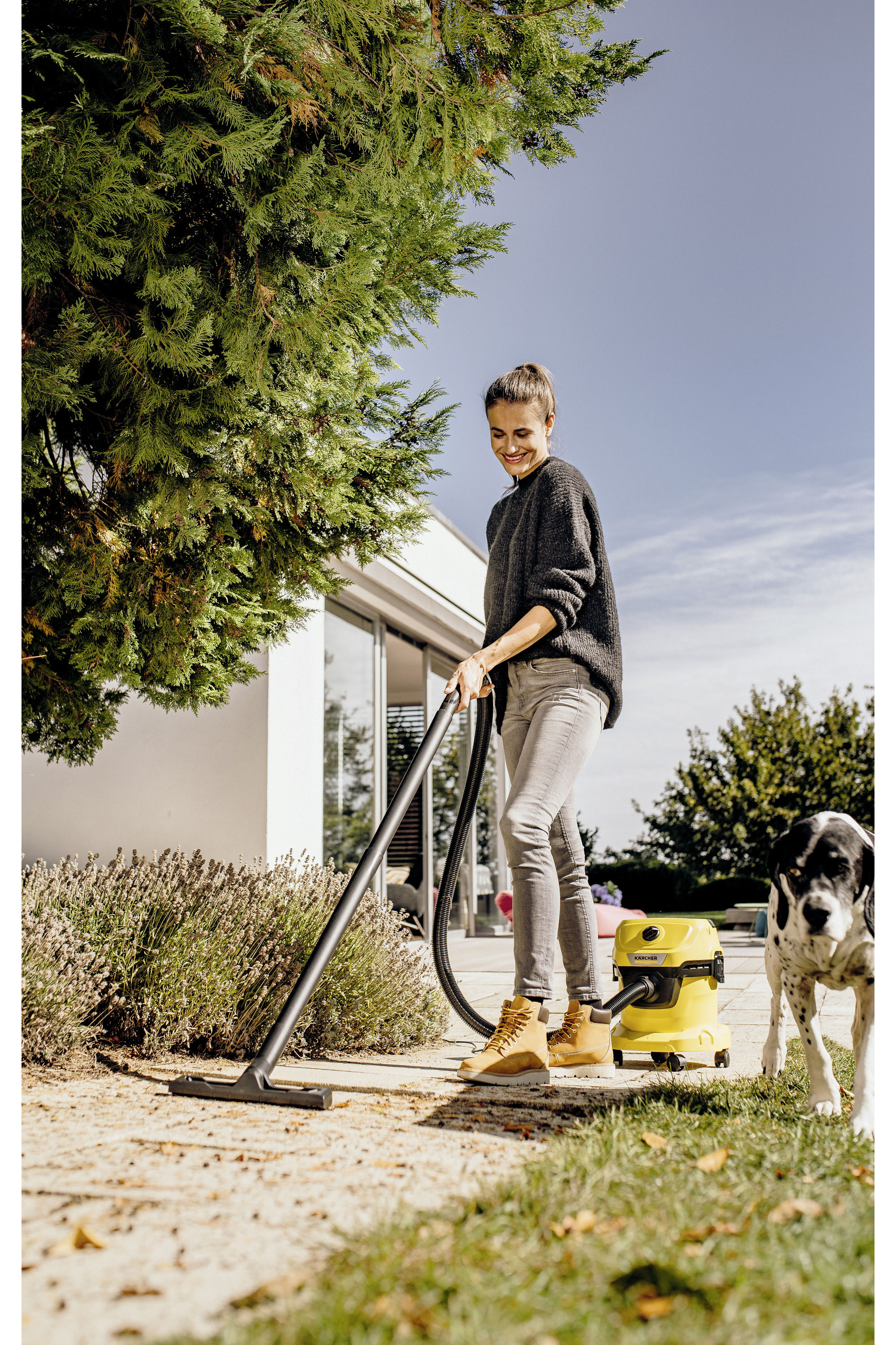 Kobieta odkurza żółtym odkurzaczem Kärcher w ogrodzie, pies patrzy.