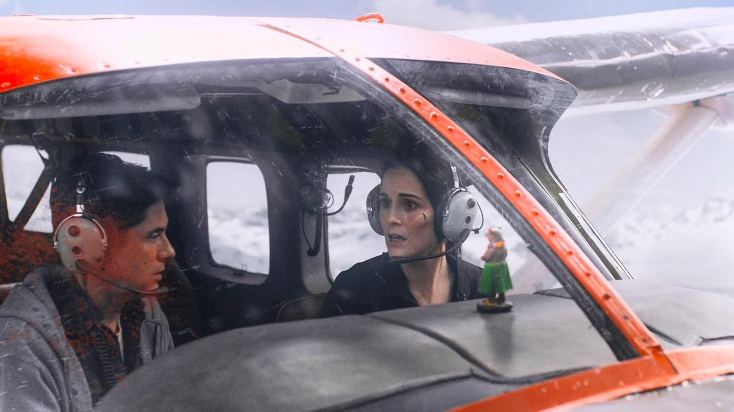 Zwei Personen in einem Flugzeug, eine schaut besorgt, der Pilot steuert. Schnee draußen.