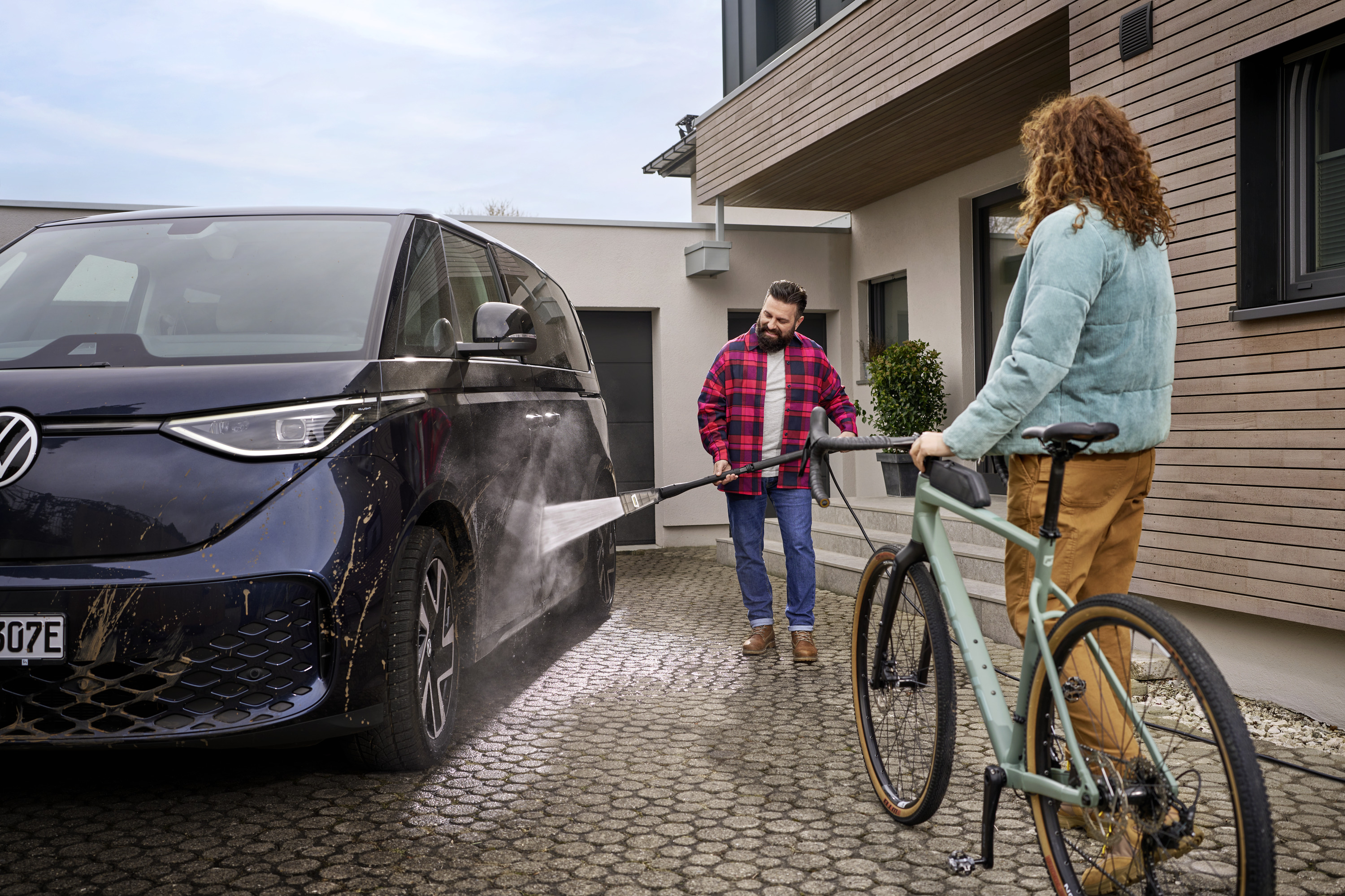 Mężczyzna myje niebieski samochód myjką ciśnieniową, a obok stoi kobieta z rowerem.