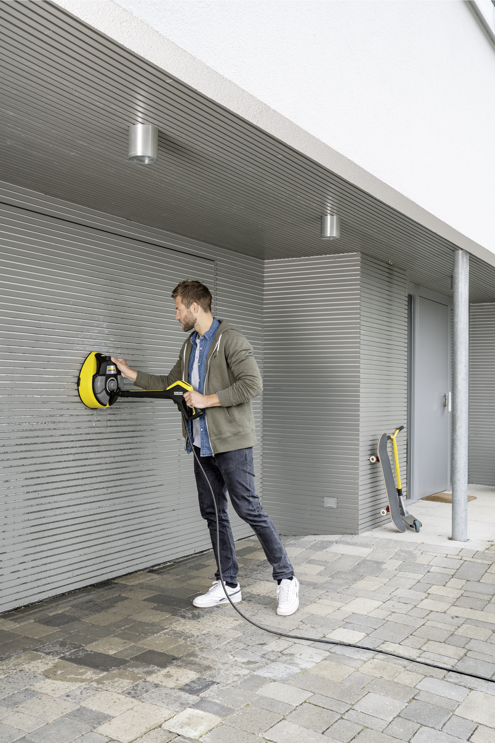 Mężczyzna czyszczący szarą ścianę żółto-czarnym urządzeniem Kärcher; w tle skuter.