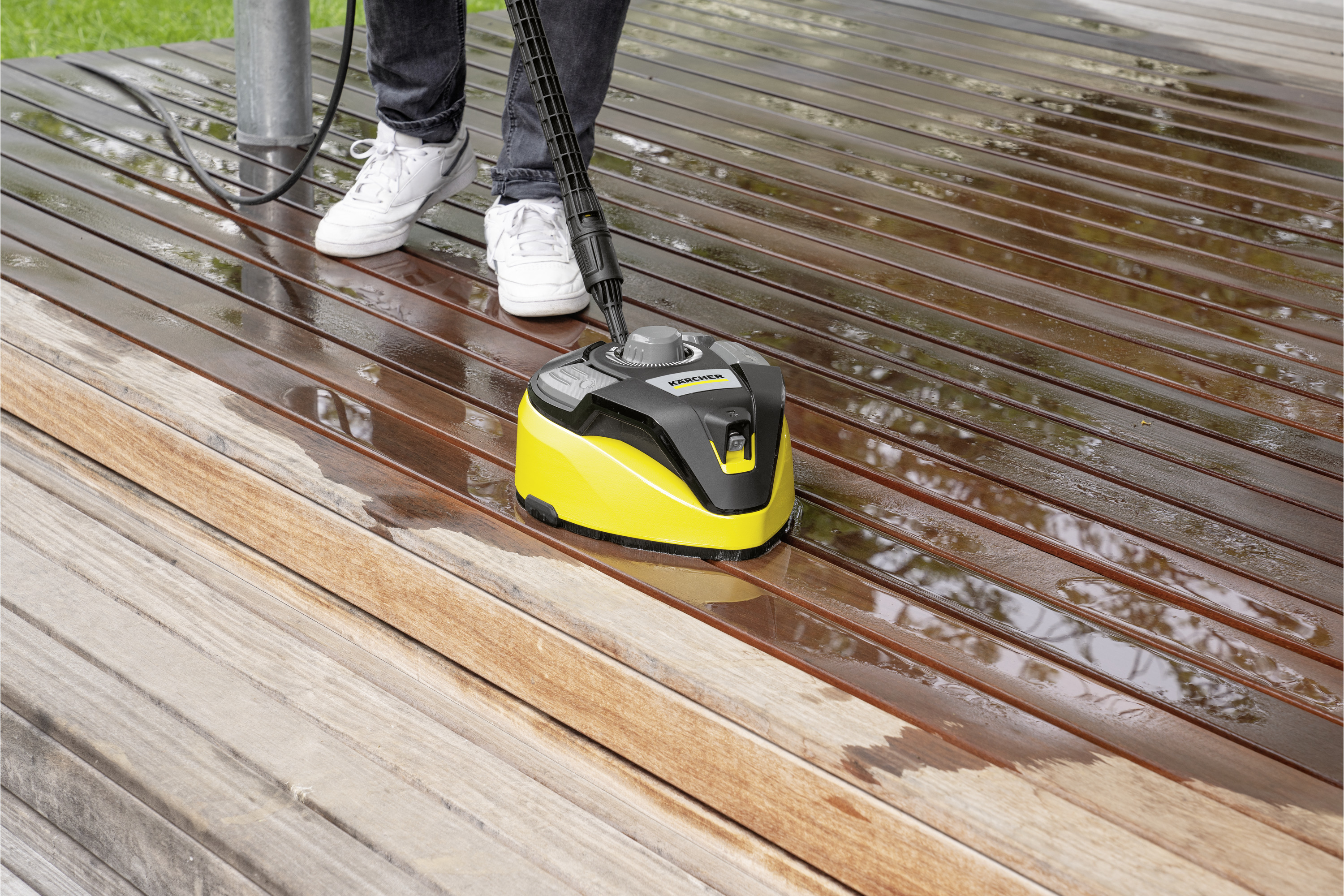 Osoba czyszcząca drewniany taras żółto-czarnym urządzeniem Kärcher; widoczne stopy.