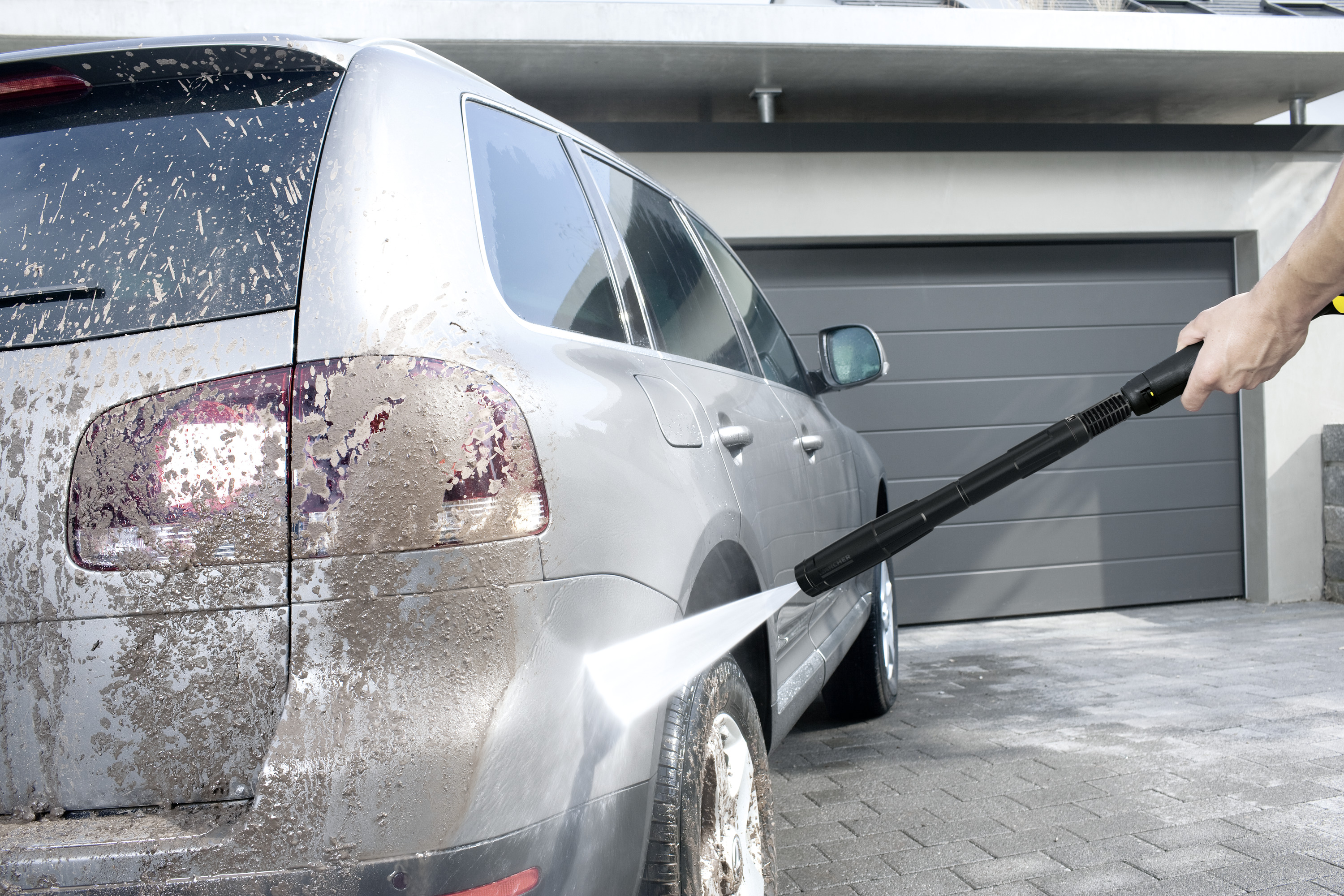 Osoba czyści brudny samochód myjką ciśnieniową Kärcher, szary samochód przed garażem.