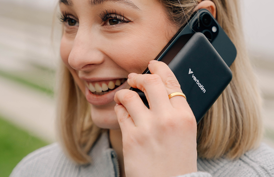 Mosolygó nő telefonál egy fekete töltővel, zöld táj a háttérben.