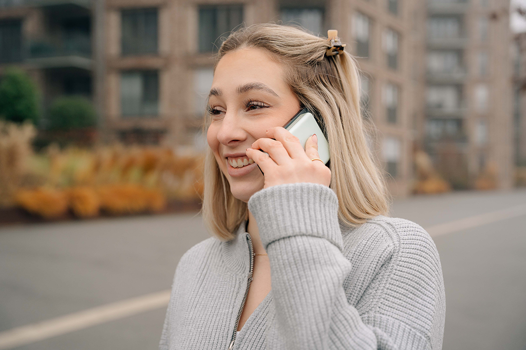 Egy mosolygó nő telefonál. Egy épület előtt a szabadban van, a háttérben növényekkel.