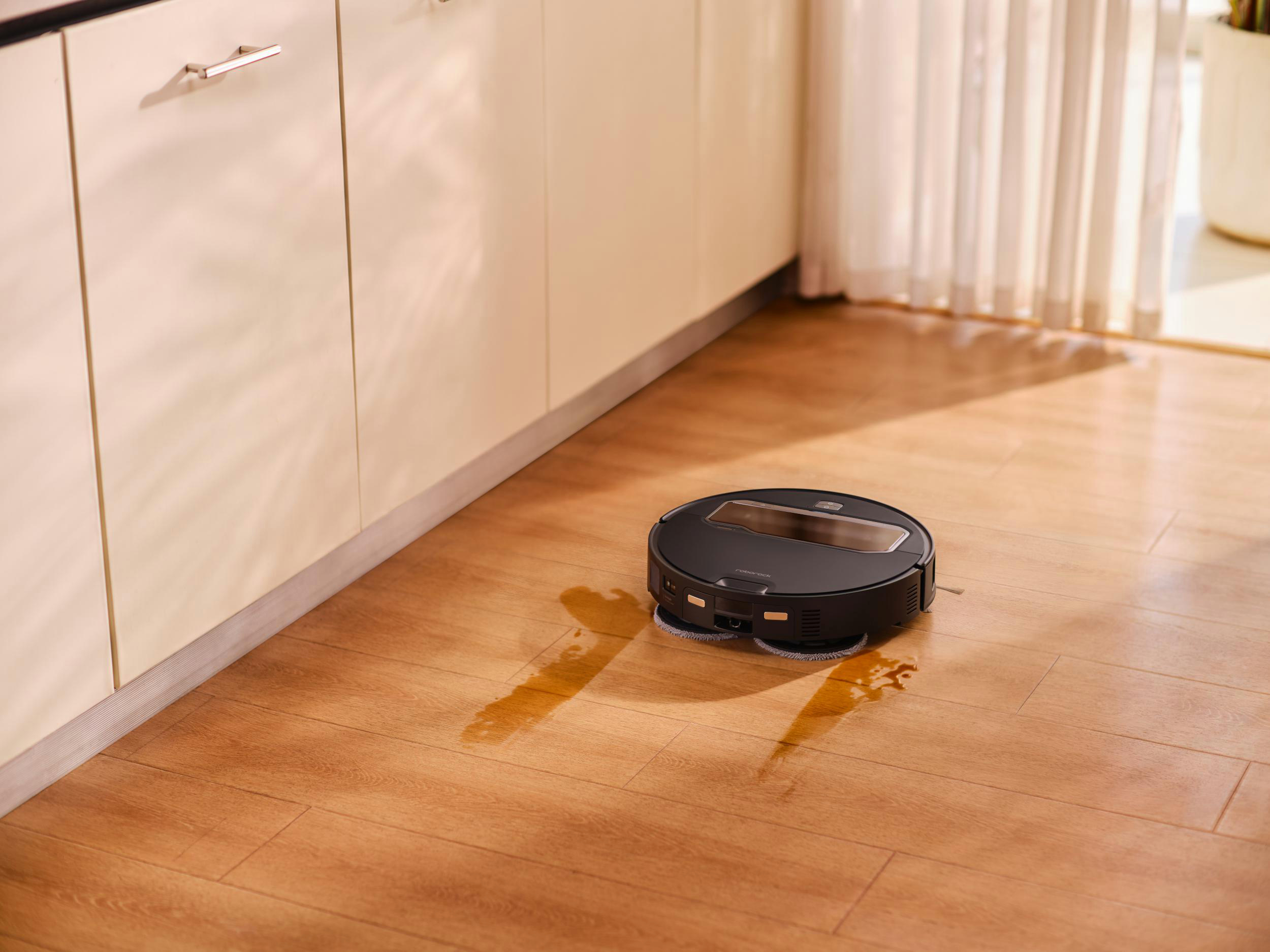 Ein schwarzer Roboter-Staubsauger reinigt einen Holzboden. Es gibt Wasserflecken.