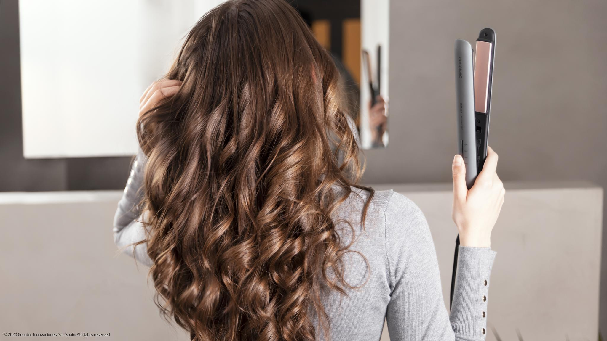Kobieta trzyma szarą prostownicę i patrzy na swoje kręcone włosy. Napis głosi: Cecotec, Bamba RitualCare.