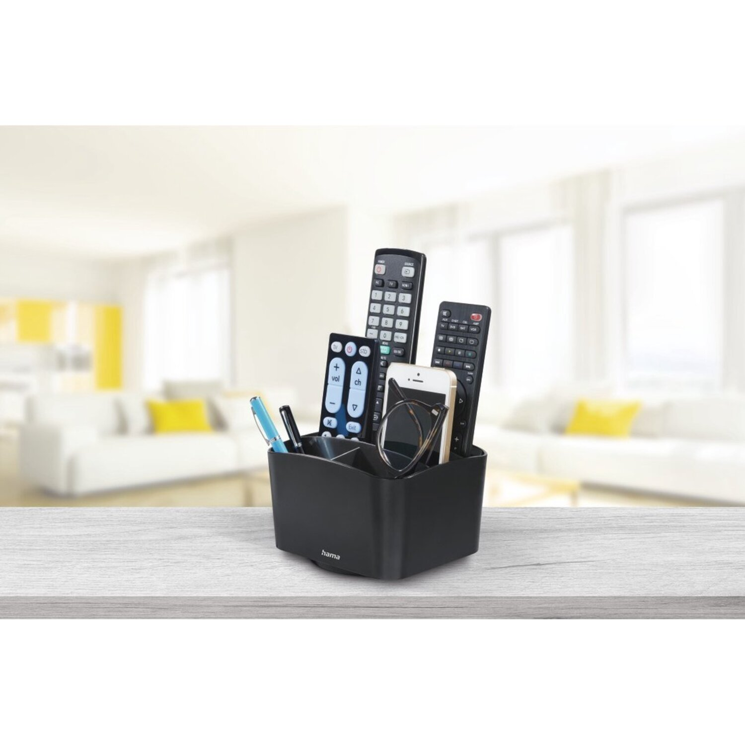 Black organizer on a wooden table, holding remotes, pens, glasses, and a phone. Living room background.