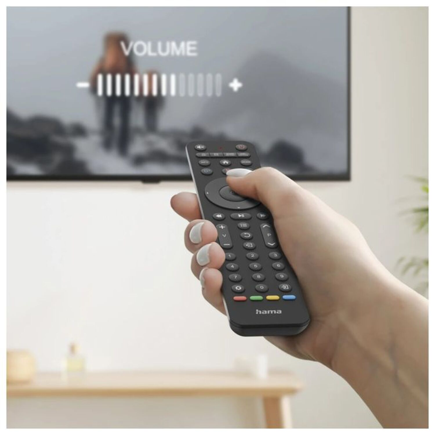 Hand holding a black remote control adjusting volume on a TV. The TV shows a blurred scene of people.