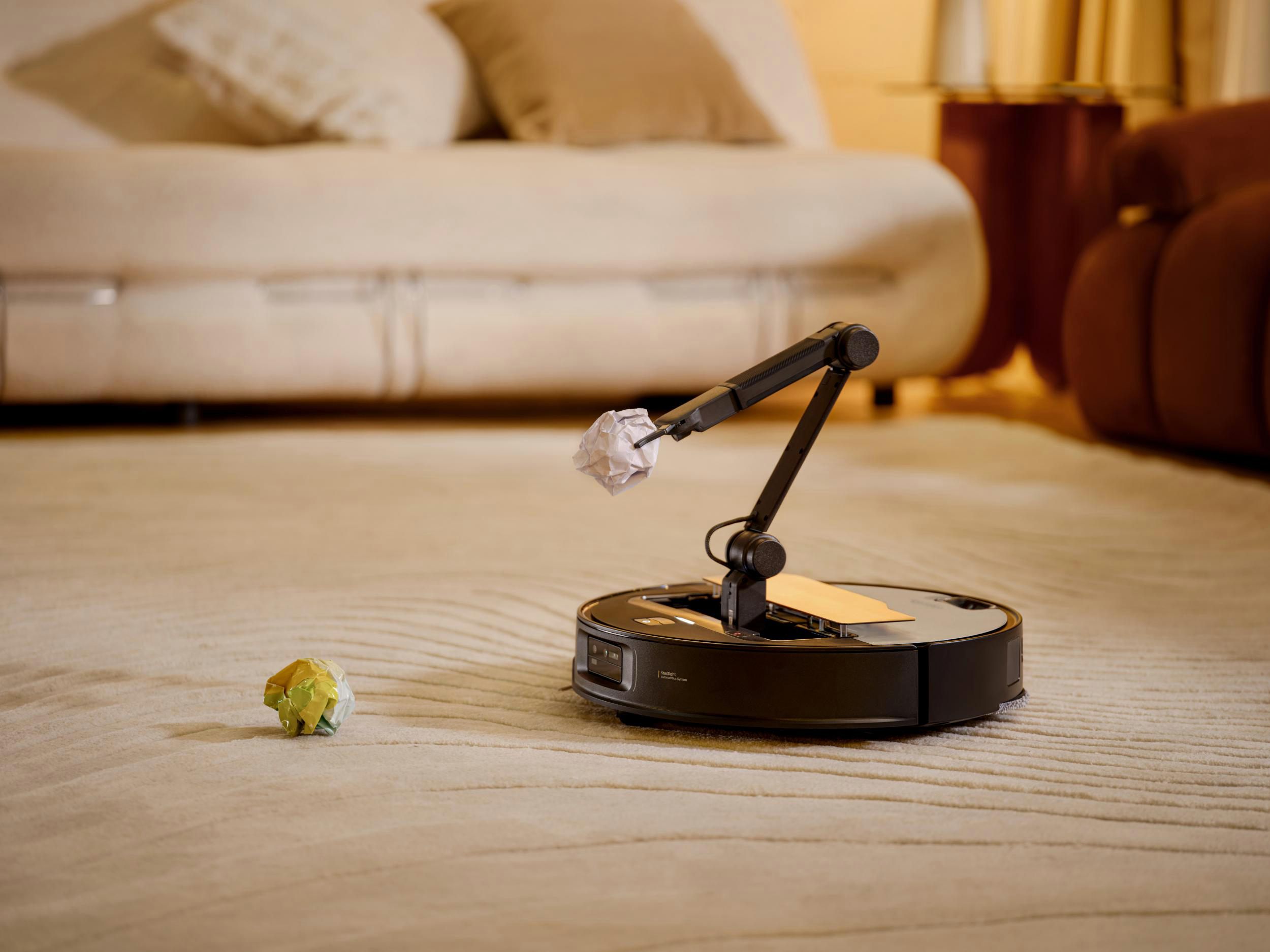 Roboterarm hebt Papierknäuel. Hintergrund: Teppich, Möbel.