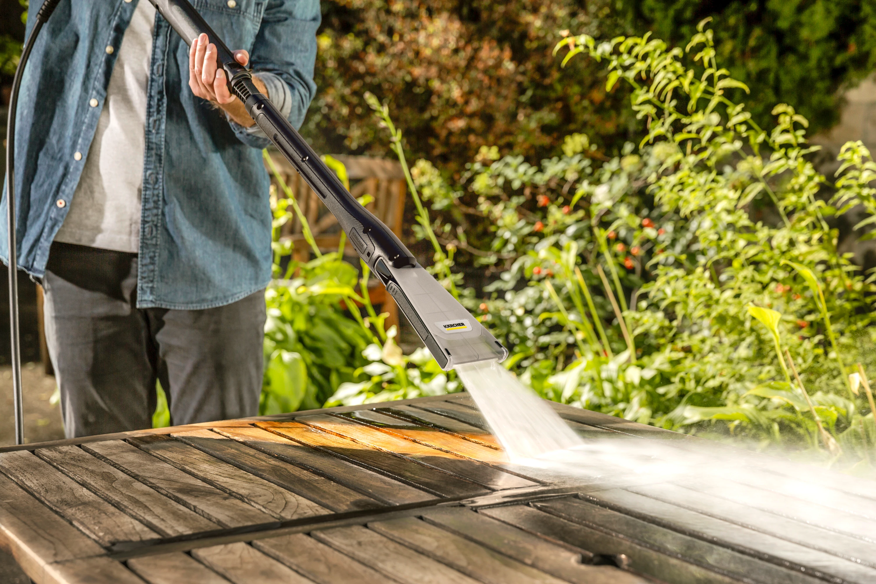 Osoba czyści drewniany stół myjką ciśnieniową. Woda rozpryskuje się na stole.